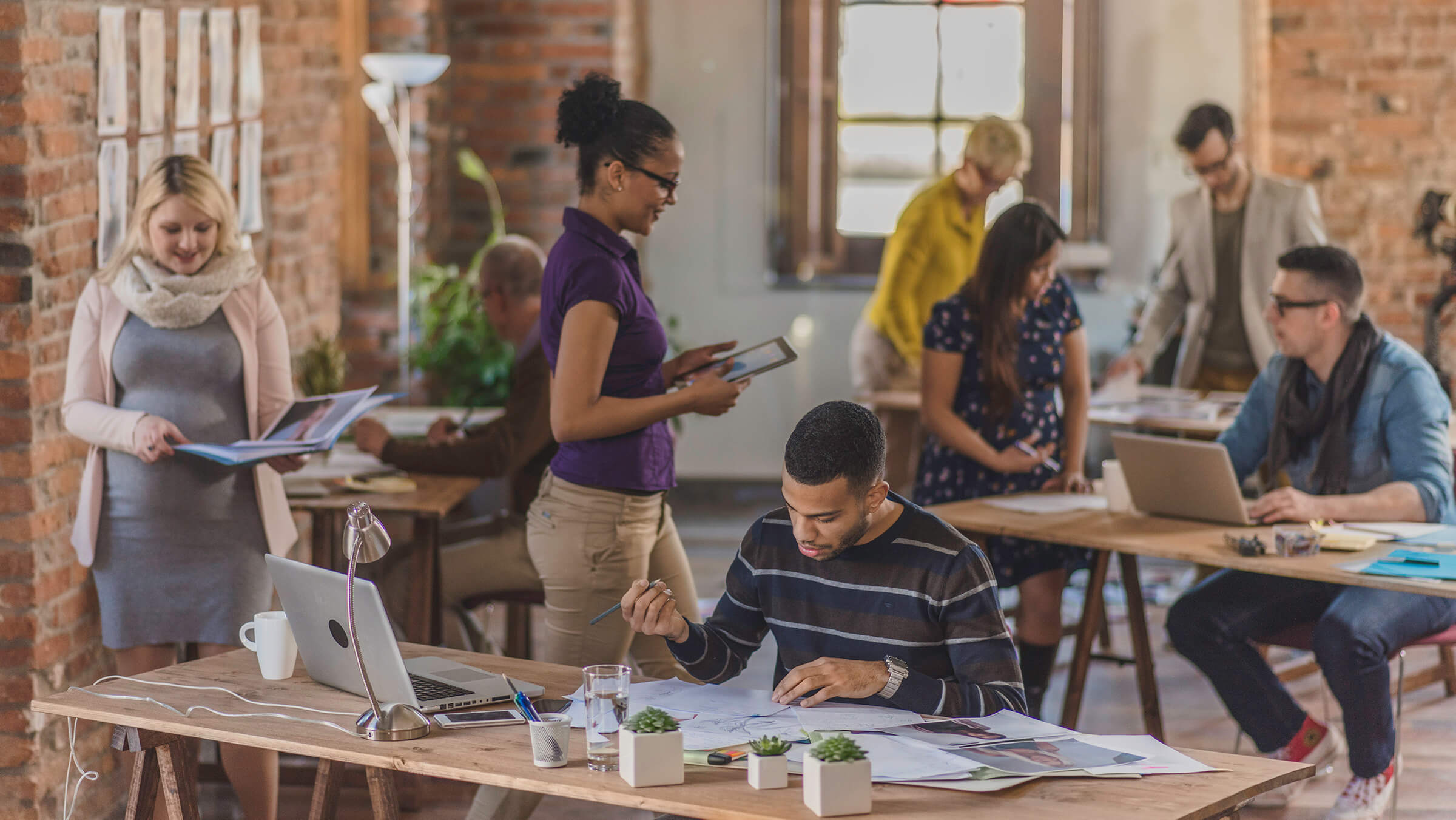 collaborative workspace with diverse professionals discussing projects, engaged in teamwork, and using laptops, modern office with brick walls and natural light